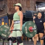 Seorang wanita sedang merekam video di area gym dengan latar belakang peralatan angkat beban profesional.