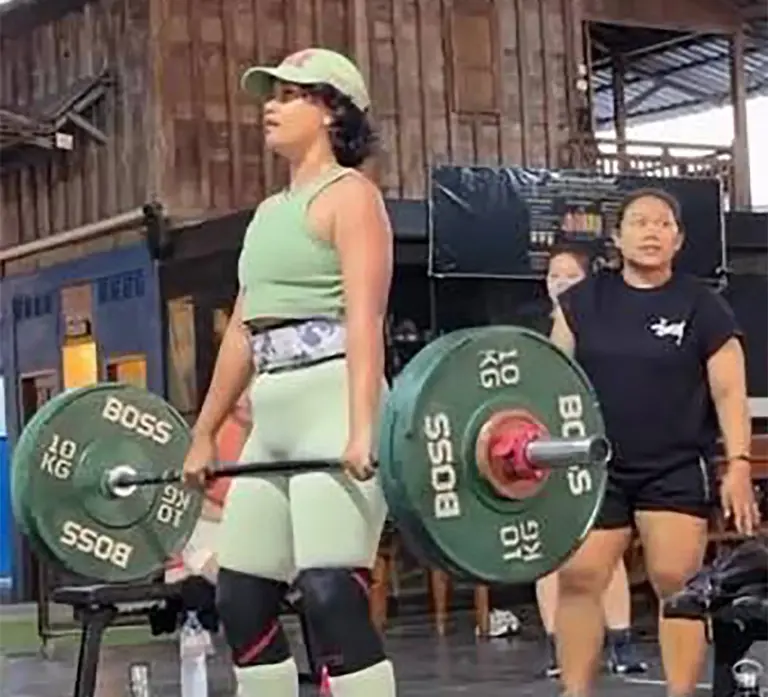 Seorang wanita sedang merekam video di area gym dengan latar belakang peralatan angkat beban profesional.