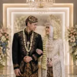 Pasangan pengantin mengenakan perpaduan busana adat Jawa dan Sunda di pelaminan modern minimalis.